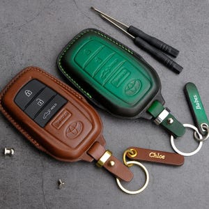 May include: Two leather keychains with personalized name tags. One keychain is green with a black leather key fob and the other is brown with a brown leather key fob. Both keychains have metal rings and a metal hook.