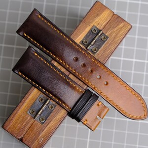 May include: Two brown leather watch straps with contrasting orange stitching. The straps are laid out on a wooden box with a metal hinge.