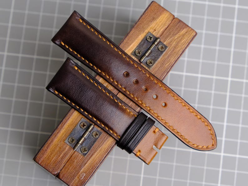 May include: Two brown leather watch straps with contrasting orange stitching. The straps are laid out on a wooden box with a metal hinge.