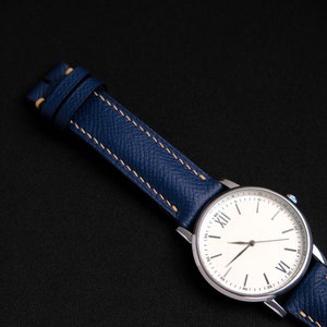 May include: A silver wristwatch with a white face and Roman numeral hour markers. The watch has a blue leather band with light brown stitching.