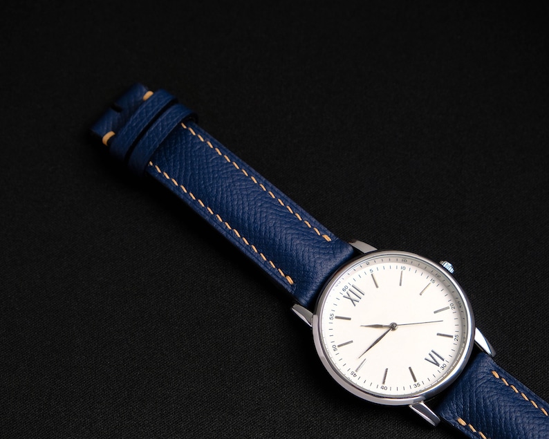 May include: A silver wristwatch with a white face and Roman numeral hour markers. The watch has a blue leather band with light brown stitching.