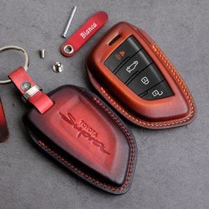 May include: A red and black leather key fob with the word "Supra" and "Toyota" embossed on it. The key fob has a silver ring and a red leather strap. A separate brown leather key fob with a silver key with buttons is also shown.