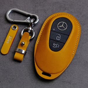 May include: A yellow leather key fob with a black button and a silver Mercedes-Benz logo. The key fob is attached to a silver key ring with a yellow leather strap and a silver tag that says "Ellen".
