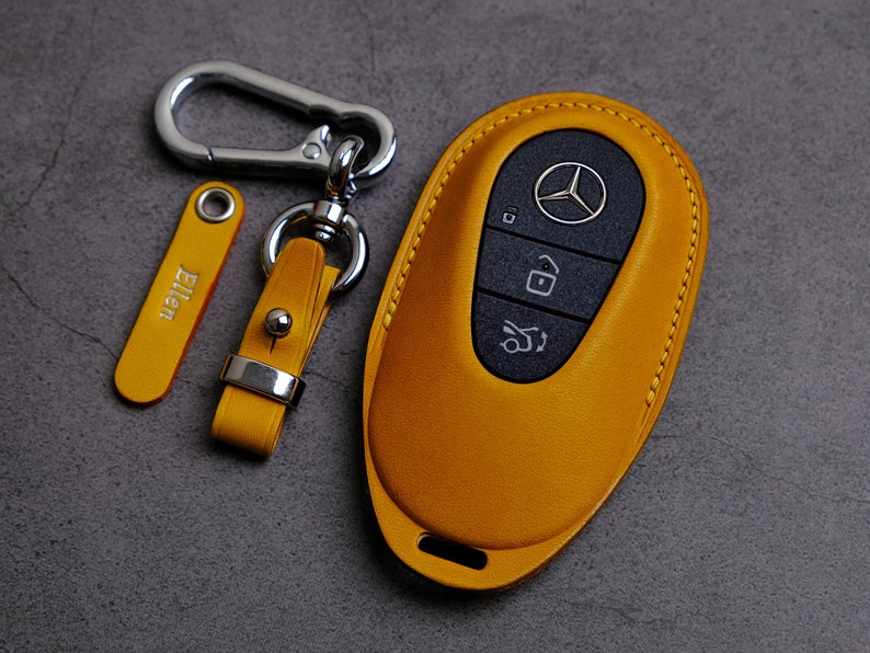 May include: A yellow leather key fob with a black button and a silver Mercedes-Benz logo. The key fob is attached to a silver key ring with a yellow leather strap and a silver tag that says "Ellen".
