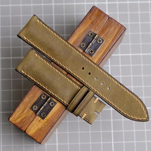 May include: Olive green leather watch strap with cream stitching, resting on a small wooden box with metal hinges. The strap has multiple holes for adjustment and a buckle. The background is a gray grid.