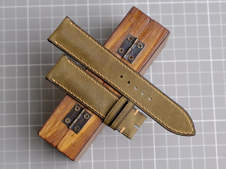 May include: Olive green leather watch strap with cream stitching, resting on a small wooden box with metal hinges. The strap has multiple holes for adjustment and a buckle. The background is a gray grid.