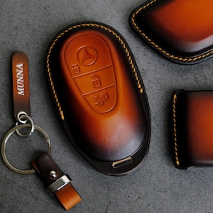 May include: A leather key fob and key ring set in shades of brown and tan. The key fob has embossed buttons and a car logo. The key ring has the name "MUNNA" printed on the leather strap. The set is stitched with yellow thread.