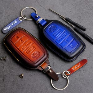 May include: Two leather keychains with personalized name tags. One keychain is brown with a blue and white car key design. The other keychain is blue with a black and white car key design. Both keychains have silver key rings.