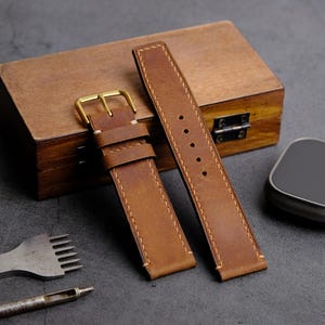 May include: Two brown leather watch straps with gold buckles and stitching. The straps are laid out on a wooden box and a gray surface.