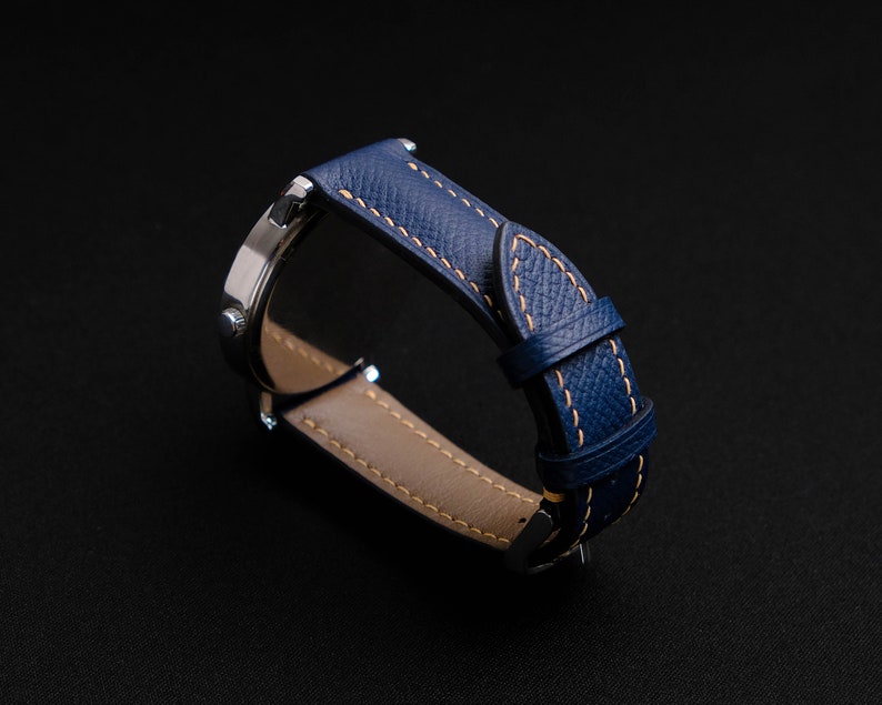 May include: A blue leather watch band with a light brown stitching detail. The band is shown from the side, with the watch face not visible.