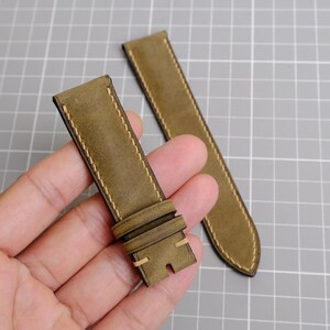 May include: Two olive green watch straps with cream-colored stitching. One strap is held in a hand, showing the buckle and the other strap is lying on a gray surface. The straps are made of a textured material.