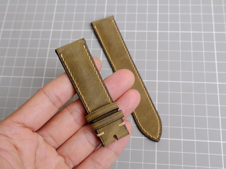 May include: Two olive green watch straps with cream-colored stitching. One strap is held in a hand, showing the buckle and the other strap is lying on a gray surface. The straps are made of a textured material.