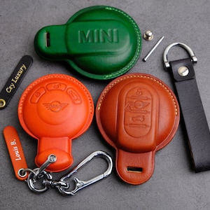Può includere: Custodie per portachiavi in pelle verde, arancione e marrone, con logo MINI in rilievo. Include un portachiavi in pelle nera e un moschettone argentato. Un portachiavi ha il testo "Cey Luxury" e un altro "Louis B."