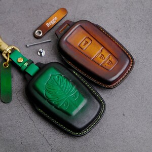 May include: Two leather key covers. One is brown with a green leaf design and the other is green with a brown leaf design. Both have stitching around the edges.
