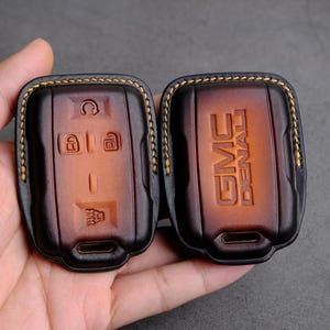 Puede incluir: Dos llaveros de cuero marrón con costuras doradas. Un llavero tiene el texto "GMC DENALI" grabado en relieve en la parte delantera. El otro llavero tiene una serie de botones y símbolos grabados en relieve en la parte delantera.