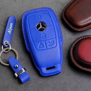 May include: A blue leather key fob with a Mercedes-Benz logo and four buttons. The key fob is on a gray surface.