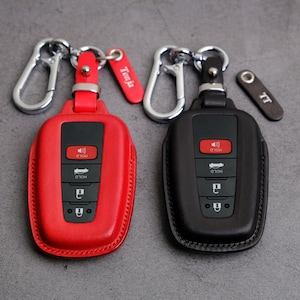 May include: Two key fobs with leather key covers. One is red and the other is black. Both have a silver key ring with a metal loop. The red key fob has a tag that says "Tesla". The black key fob has a tag that says "TT".