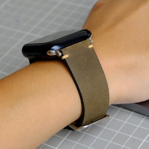 Peut inclure: Une montre intelligente noire avec un bracelet en cuir vert est portée au poignet. Le bracelet a un design simple avec des coutures de couleur claire et une boucle argentée. La montre est sur une surface quadrillée grise.