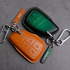 Puede incluir: Dos fundas de llavero de cuero en naranja y verde, con mosquetones plateados. El llavero naranja tiene el nombre "Neill" en una pequeña etiqueta. El llavero verde tiene el nombre "Rosemary" en una pequeña etiqueta. Ambos tienen diseños de botones de llave.