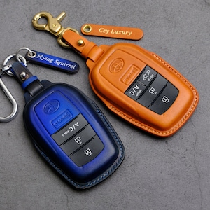 May include: Two leather key fobs for a car key. The key fobs are in blue and orange leather. The blue key fob has the text "HOLD A/C HOLD" and the orange key fob has the text "HOLD A/C HOLD". Both key fobs have a silver metal ring.