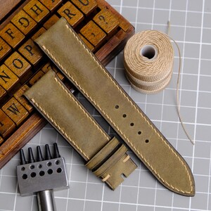 May include: Olive green leather watch straps with cream stitching, alongside a spool of twine, a wooden letter set, and a metal punch tool. The watch straps are new and ready to be installed on a watch.