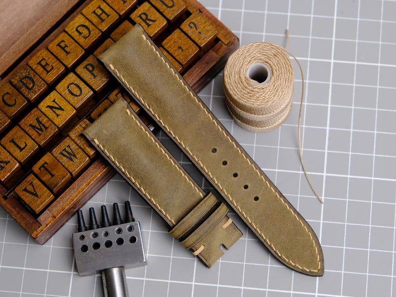 May include: Olive green leather watch straps with cream stitching, alongside a spool of twine, a wooden letter set, and a metal punch tool. The watch straps are new and ready to be installed on a watch.