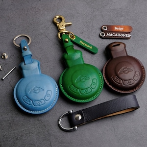 Può includere: Tre portachiavi in ​​pelle di diversi colori, ognuno con un piccolo pulsante rotondo con un logo. I portachiavi sono etichettati "Badge", "MACAIONE29" e "Byxmark".