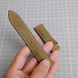 May include: Olive green leather watch strap with cream stitching. The strap has several holes for adjustment and a buckle. The watch band is shown on a gray grid background.