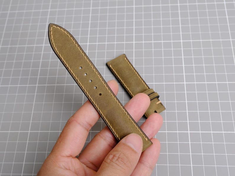 May include: Olive green leather watch strap with cream stitching. The strap has several holes for adjustment and a buckle. The watch band is shown on a gray grid background.