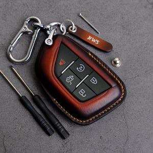 Puede incluir: Un llavero de cuero marrón con costuras en los bordes. El llavero tiene cinco botones con símbolos en ellos. El llavero está unido a un llavero plateado con un gancho plateado.