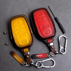 May include: Two leather car key fobs, one yellow and one red, with a Toyota logo. The yellow fob has the word "GOD" on a small tag, and the red fob has the name "Alisha". A screwdriver and two silver carabiner clips are also present.