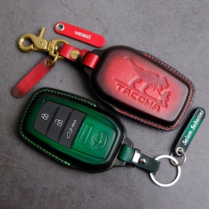Puede incluir: Dos llaveros de cuero. Uno es rojo con "TACOMA" en relieve y un cierre dorado. El otro es verde con un panel de llaves negro. También hay una etiqueta roja con "wasazi" y una etiqueta verde con "Salem Sabatini".