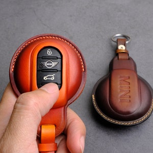 Pode incluir: Um chaveiro redondo de couro marrom com uma borda costurada. O chaveiro tem um anel de metal e um pequeno botão de metal. O chaveiro é projetado para segurar uma chave de carro e tem a palavra "MINI" gravada em relevo.