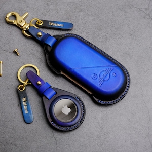 Può includere: Due portachiavi in ​​cuoio blu con accessori dorati. Un portachiavi ha un portachiavi rotondo in cuoio blu con un piccolo logo Apple bianco. L'altro portachiavi ha un portachiavi rettangolare in cuoio blu con la parola "Wattson" impressa in lettere dorate. Entrambi i portachiavi hanno un anello dorato e un gancio dorato.