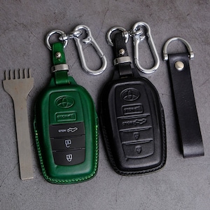 Op de afbeelding: Twee leren sleutelhangers met metalen sleutelringen. Een hanger is groen en de andere is zwart. Beide hangers hebben een auto-sleutelontwerp met het woord "HOLD" en een auto-icoon.