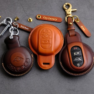 Può includere: Tre copri chiavi in pelle per un portachiavi per auto. Le custodie sono marroni e beige con cuciture sui bordi. Le custodie hanno la parola "Mini" impressa su di esse. Le custodie sono attaccate agli anelli portachiavi con anelli metallici. Gli anelli portachiavi hanno etichette con le parole "Julchen", "Cey Luxury" e "Kika" su di essi.