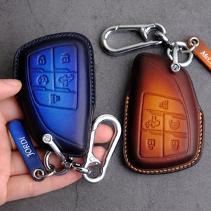 May include: Two leather key fobs with metal key rings and a silver metal clasp. The key fob on the left is blue with the name "JORDI" on a silver tag. The key fob on the right is brown with the name "McCallum" on a brown tag.