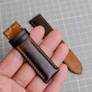 May include: Two brown leather watch straps with yellow stitching. The straps are held in a hand.