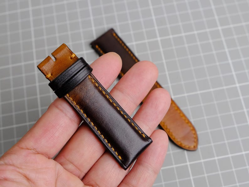 May include: Two brown leather watch straps with yellow stitching. The straps are held in a hand.