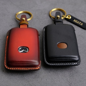 May include: Two leather keychains with gold key rings. One keychain is red with a Mazda logo and the other is black with white stitching. The black keychain has a gold tag that says "MIATA".