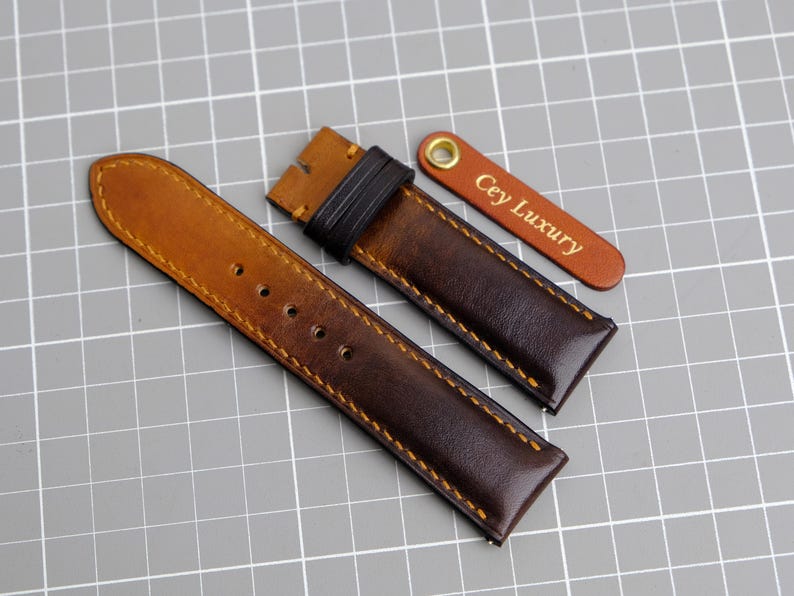 May include: Two brown leather watch straps with contrasting stitching. The strap on the left is a lighter brown and the strap on the right is a darker brown. The strap on the right has a small leather tag with the text "Cey Luxury" in gold lettering.