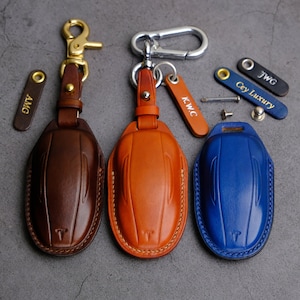 Puede incluir: Tres llaveros de cuero con cubiertas de diferentes colores: marr&oacute;n, naranja y azul. Cada llavero tiene un anillo de metal y una peque&ntilde;a etiqueta de metal con un nombre grabado. Los nombres son AMG, KWC y Cey Luxury.