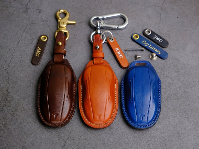 Puede incluir: Tres llaveros de cuero con cubiertas de diferentes colores: marr&oacute;n, naranja y azul. Cada llavero tiene un anillo de metal y una peque&ntilde;a etiqueta de metal con un nombre grabado. Los nombres son AMG, KWC y Cey Luxury.