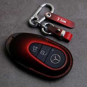 May include: A red leather key fob with a Mercedes-Benz logo and buttons. The key fob is attached to a silver key chain with a leather tag that says "J. Lim".