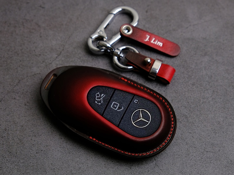 May include: A red leather key fob with a Mercedes-Benz logo and buttons. The key fob is attached to a silver key chain with a leather tag that says "J. Lim".