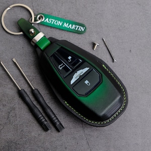May include: A green leather key fob with a black key inside. The key fob has a stitched green border and the Aston Martin logo is visible on the key. The key fob is attached to a silver key ring.