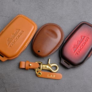 May include: Three leather keychains with the word "Tacoma" embossed on them. The keychains are brown, tan, and red. One keychain has a brass hook and a tag that says "L.A.R."
