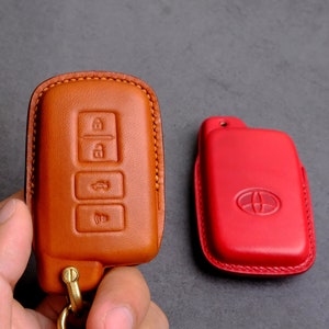 May include: Two leather key covers, one brown and one red. The brown cover has four buttons and a key ring. The red cover has a Toyota logo.