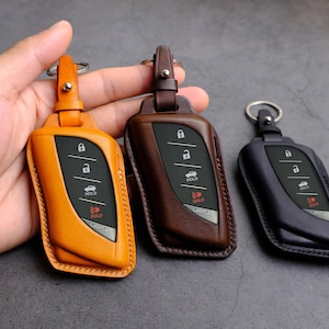 Puede incluir: Tres llaveros de cuero en diferentes colores: naranja, marrón y negro. Cada llavero tiene un lazo para llavero y un mando a distancia de entrada sin llave de cuatro botones. Los botones están etiquetados como "HOLD" y tienen un icono de coche.