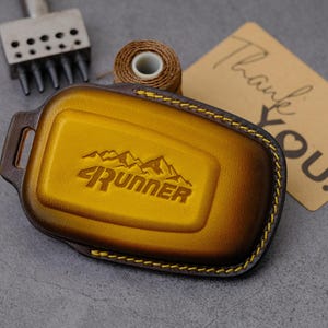 May include: A gold and brown leather key fob cover featuring an embossed mountain graphic and the text 4RUNNER. The cover is stitched with yellow thread and includes a metal hole punch tool, a spool of brown thread, and a tan thank you card.
